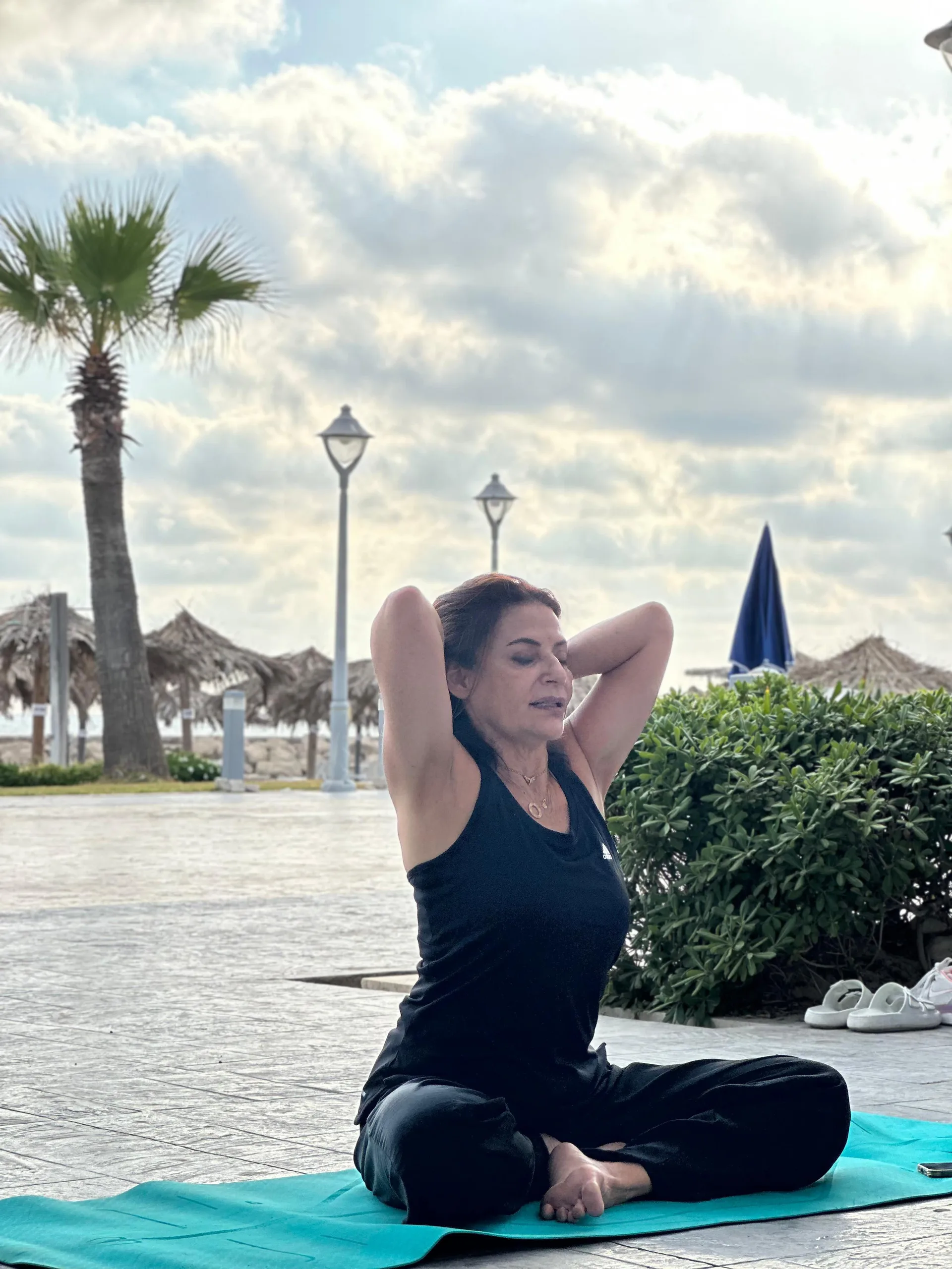 A woman in a seated yoga stretch by the sea with palm trees and beach umbrellas behind her